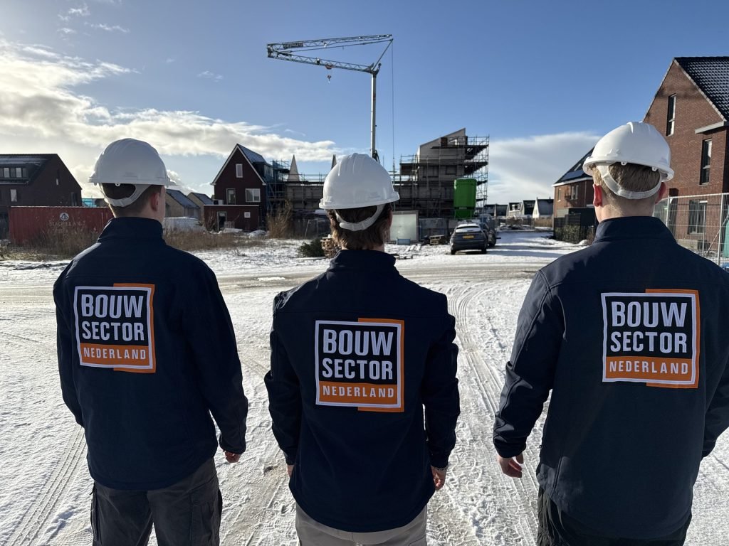 Drie collega's in werkkleding met oranje-wit logo overzien een bouwplaats met steigers